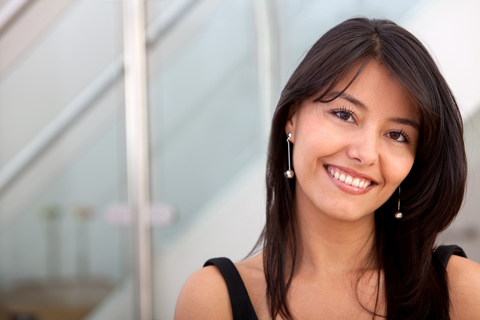 A woman smiles warmly at the camera. She has long, dark hair and is wearing drop earrings. The photo is a close-up head shot.